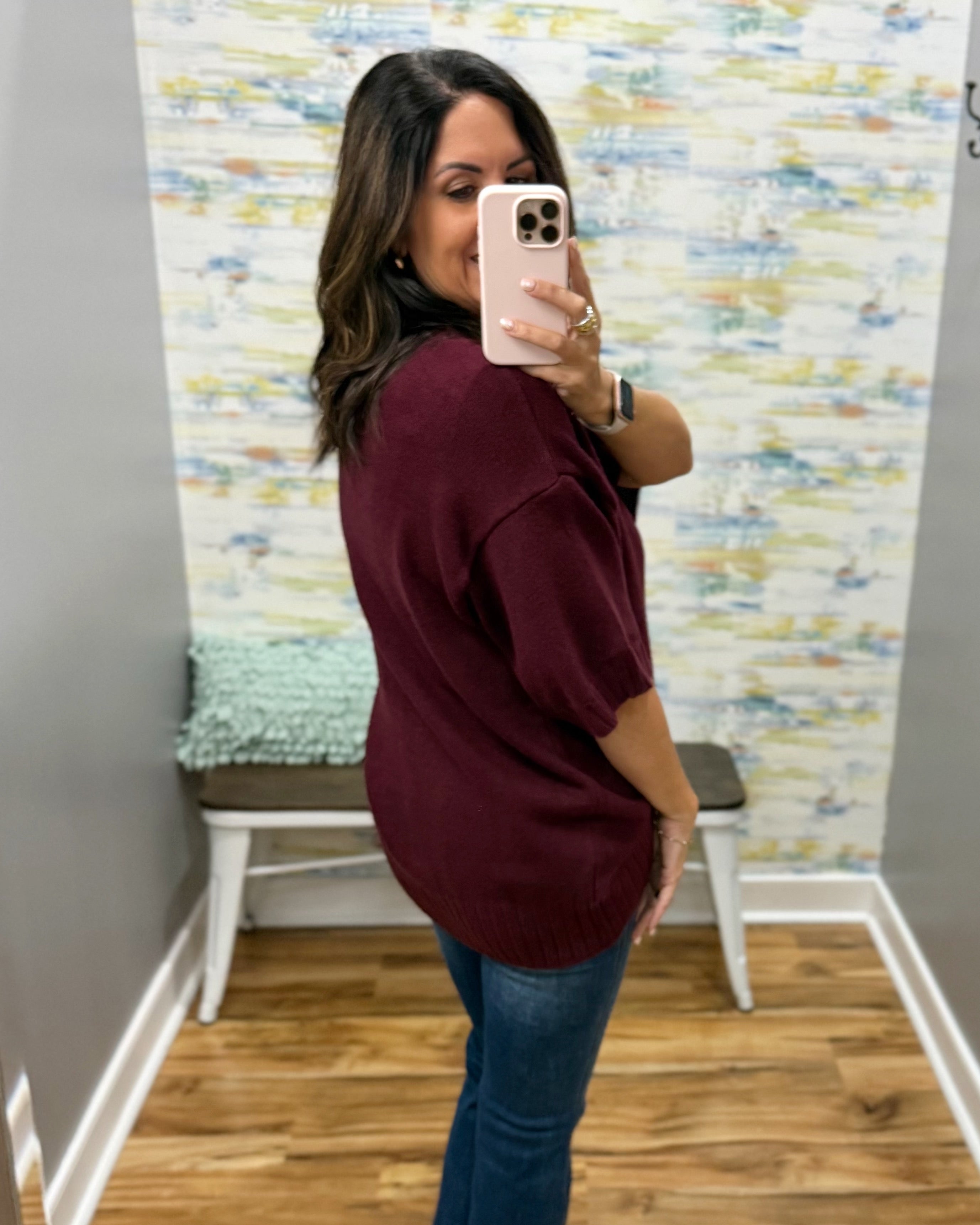 A woman taking a mirror selfie in a fitting room wearing an oversized burgundy short-sleeve knit top and blue jeans.