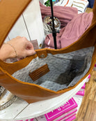 A tan leather shoulder bag with a gray and white monogram patterned interior, held open by a person's hand in a retail setting.