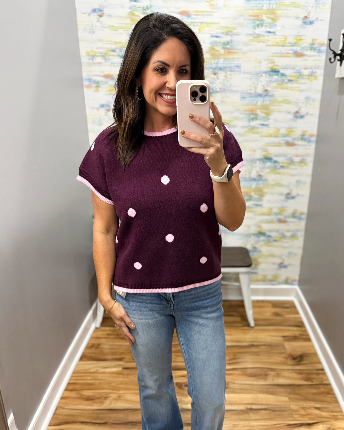A woman takes a mirror selfie wearing a short-sleeve knit top in plum with light pink trim and white polka dots, paired with light-wash blue jeans.