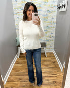 A woman takes a mirror selfie wearing a cream ribbed knit long-sleeve sweater and blue flare jeans in a fitting room with wood floors and abstract wallpaper.