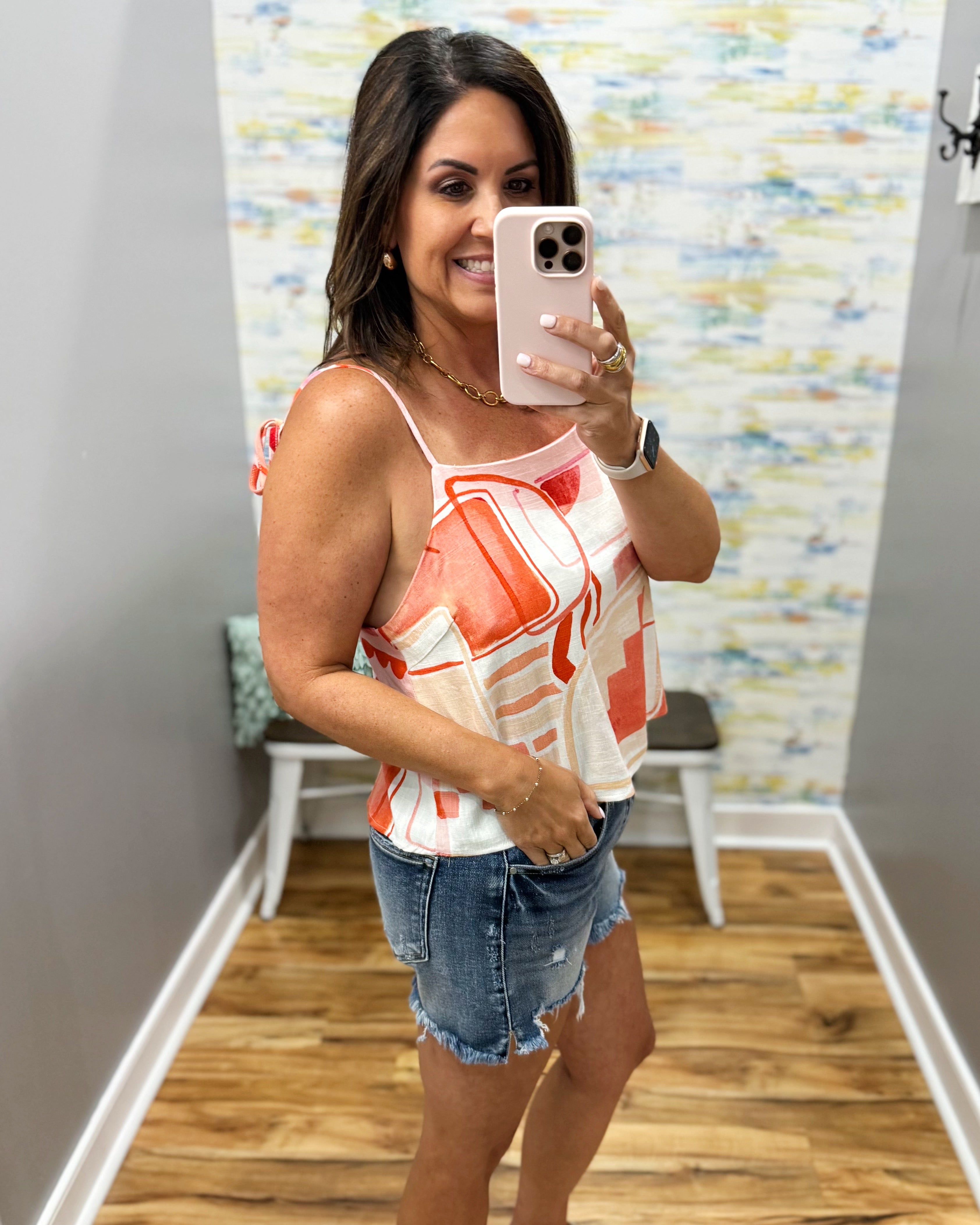 Woman taking a mirror selfie wearing a sleeveless top and denim shorts in a room with a colorful wall.