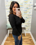 A woman taking a mirror selfie in a fitting room, wearing a black long-sleeve crewneck top and blue jeans.