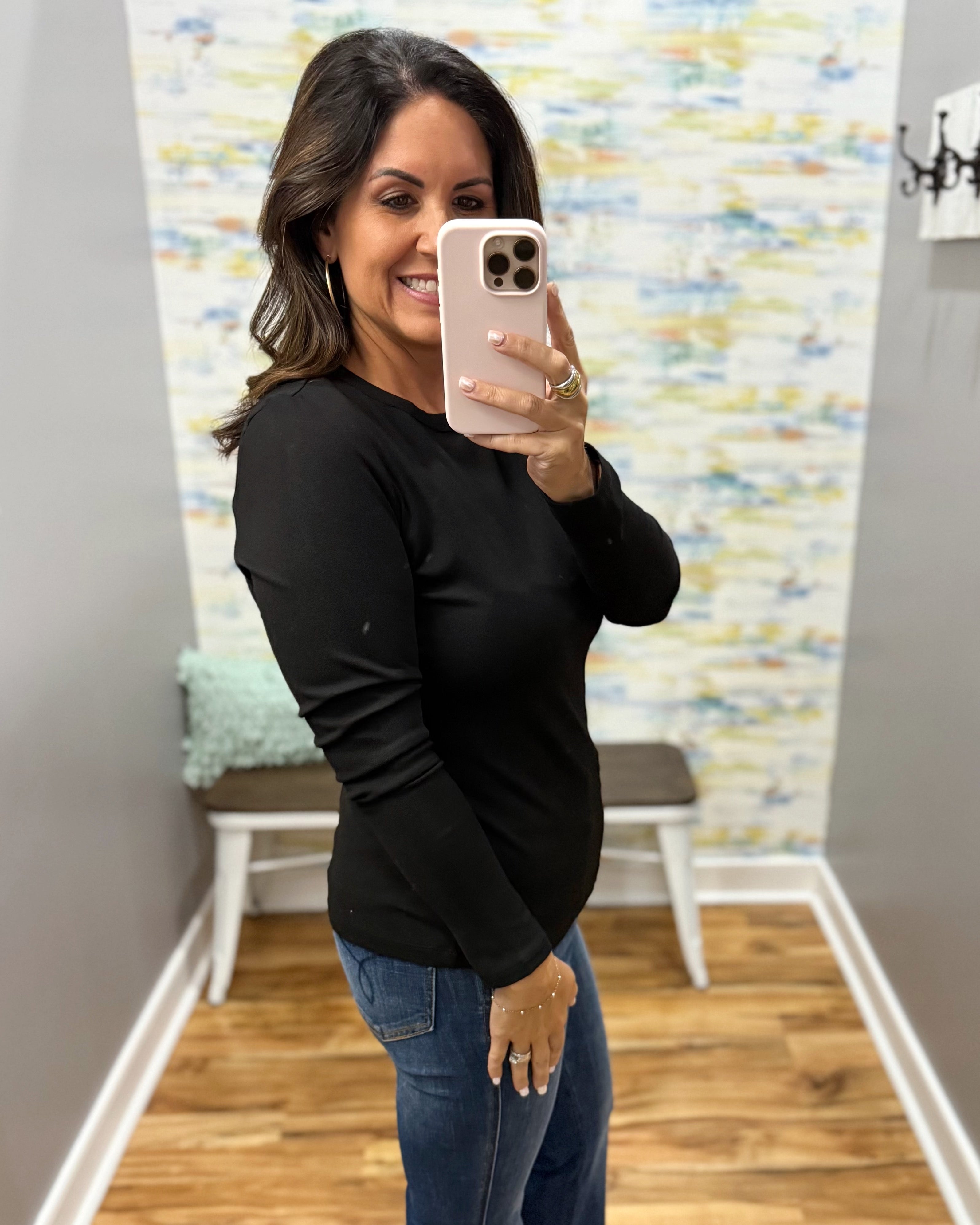 A woman taking a mirror selfie in a fitting room, wearing a black long-sleeve crewneck top and blue jeans.
