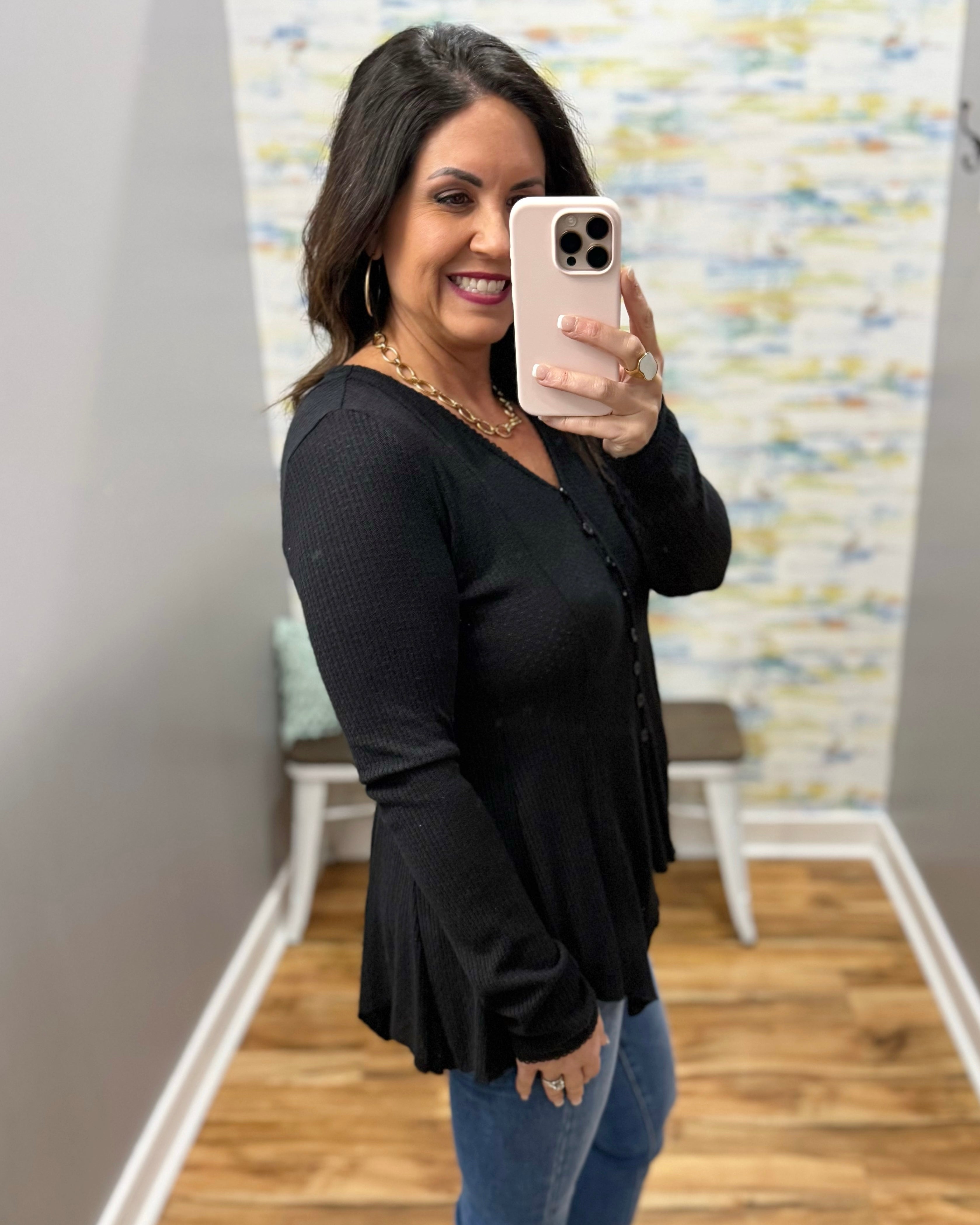Woman taking a mirror selfie wearing a black cardigan and blue jeans in a room with a patterned wall.