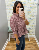 A woman takes a mirror selfie wearing a red and white gingham peplum blouse with dark blue jeans, standing in a fitting room with a patterned wall and wood floor.