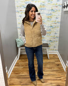 A woman takes a mirror selfie wearing a tan quilted puffer vest over a black and white striped long-sleeve shirt, dark blue jeans, and tan boots in a home fitting room.
