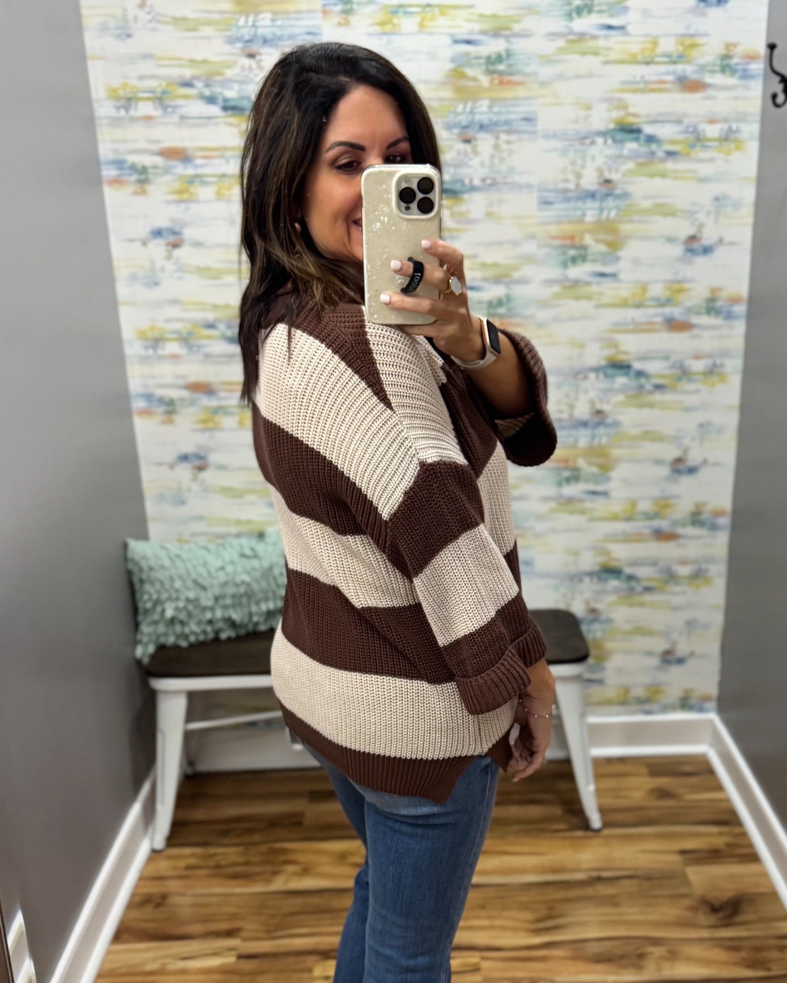 A woman takes a mirror selfie in a fitting room wearing a brown and cream striped knit sweater and blue jeans.