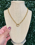 A gold chain necklace with a double-ring interlocking pendant displayed on a beige fabric jewelry bust. A hand with purple nail polish is holding the left side of the bust, and a green hedge wall serves as the background.