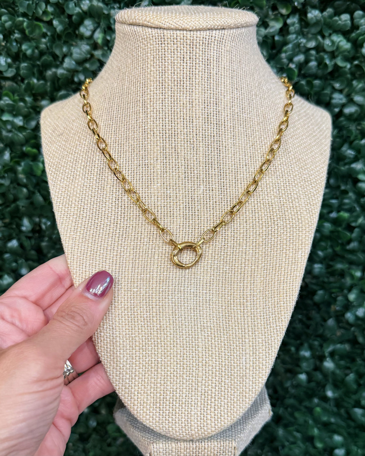 A gold chain necklace with a double-ring interlocking pendant displayed on a beige fabric jewelry bust. A hand with purple nail polish is holding the left side of the bust, and a green hedge wall serves as the background.