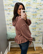 A woman takes a mirror selfie wearing a dusty rose ruffled hoodie and blue jeans in front of a patterned wallpaper wall.