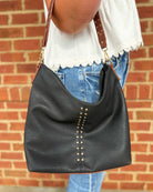 A black faux-leather slouchy hobo shoulder bag with gold-tone studs and a brown leather strap, shown being worn by a person in front of a red brick wall.