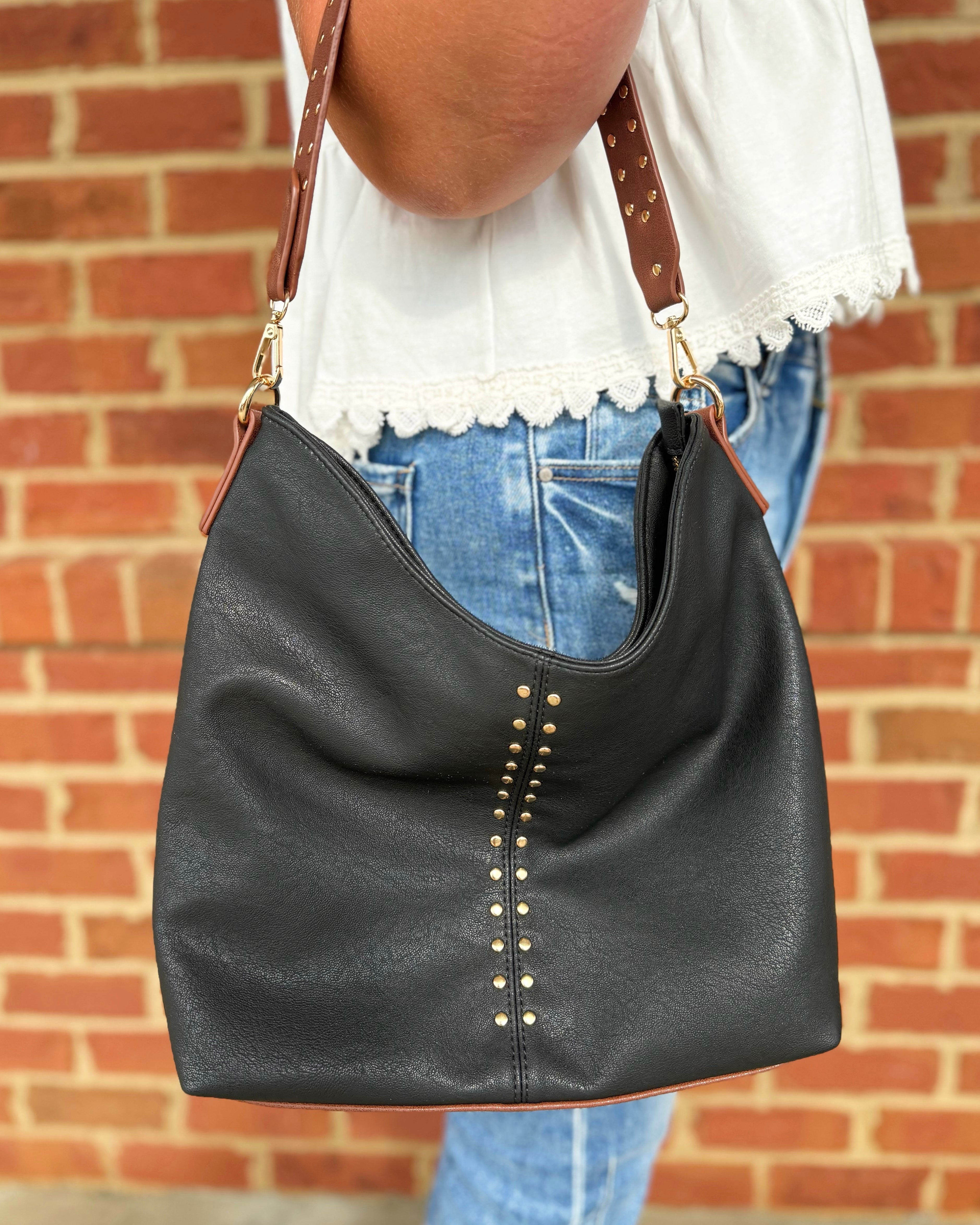 A black faux-leather slouchy hobo shoulder bag with gold-tone studs and a brown leather strap, shown being worn by a person in front of a red brick wall.