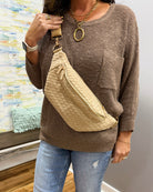 A woman wearing a brown waffle-knit sweater and light blue jeans carries a beige woven crossbody bag with a gold zipper and strap.