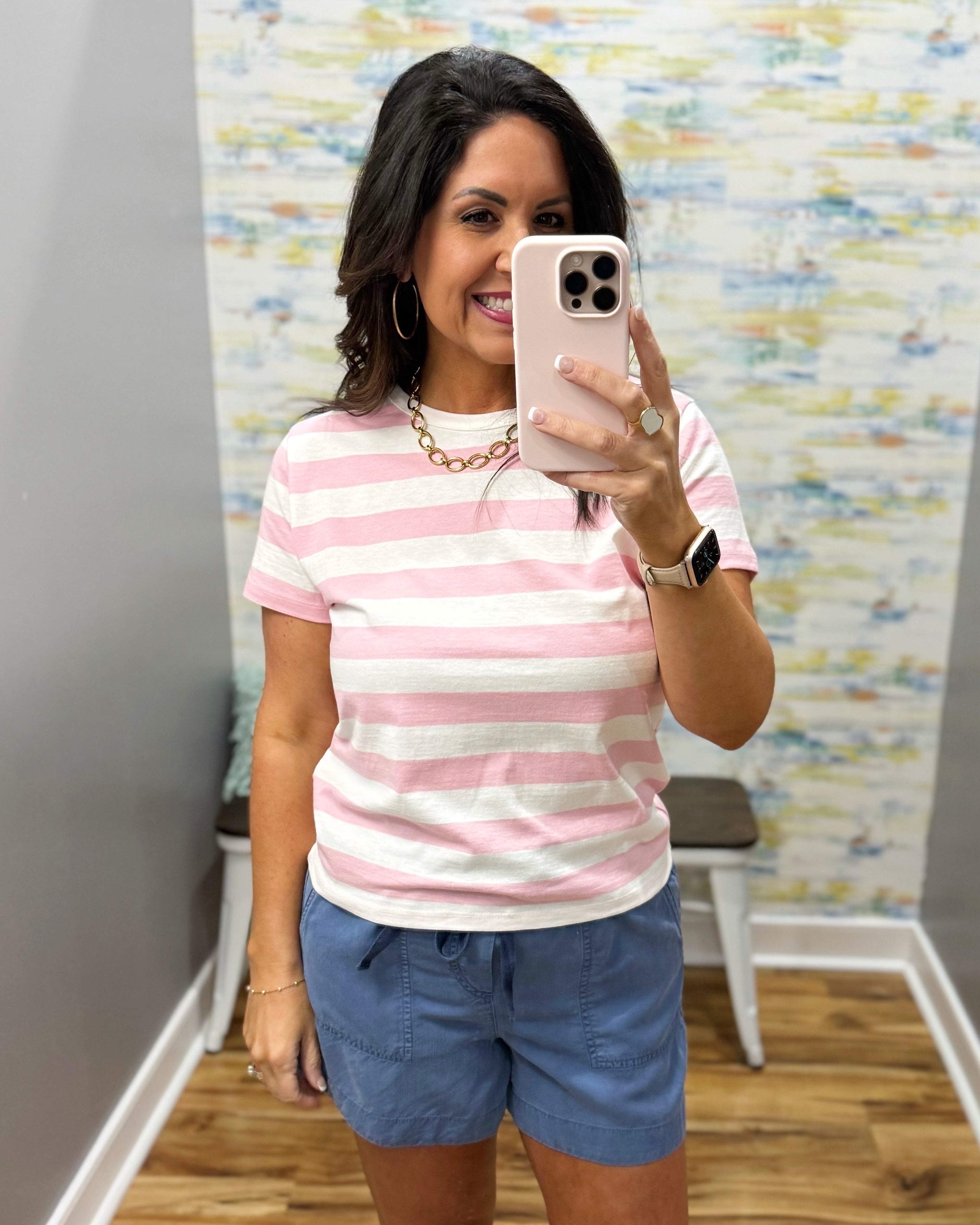 Woman taking a mirror selfie wearing a pink and white striped shirt and blue shorts.