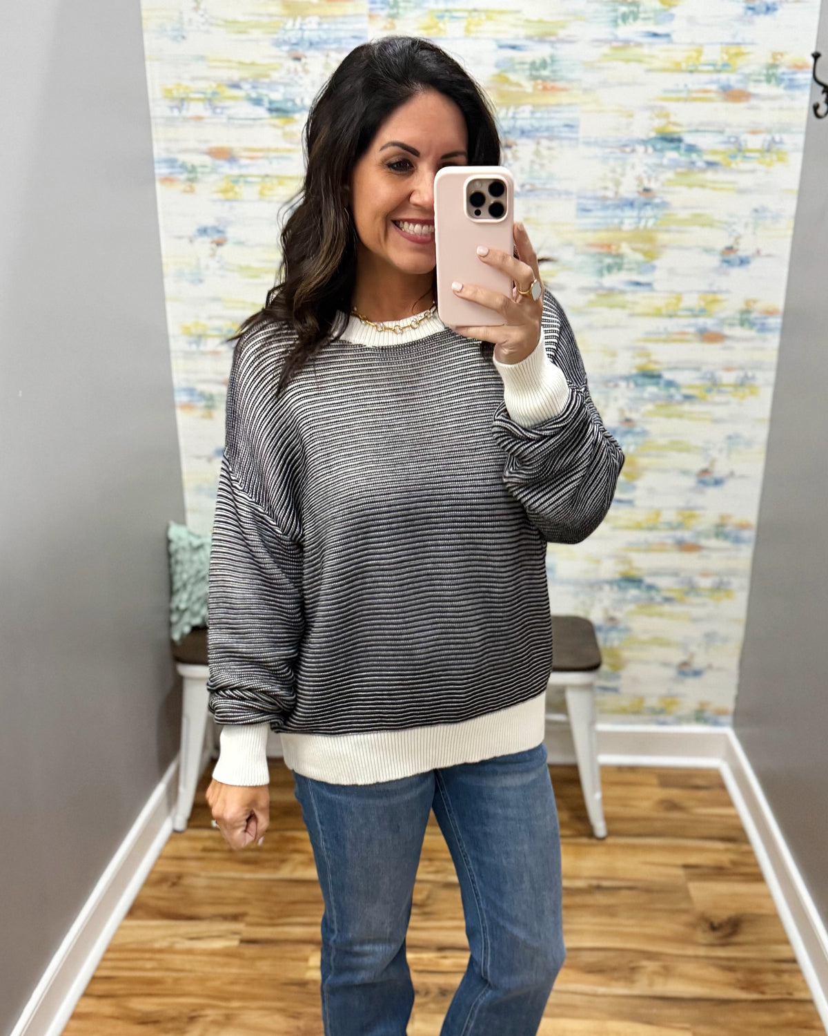 A woman takes a mirror selfie wearing a gray and white horizontally striped long-sleeve sweater with a cream ribbed hem and cuffs, paired with blue jeans.