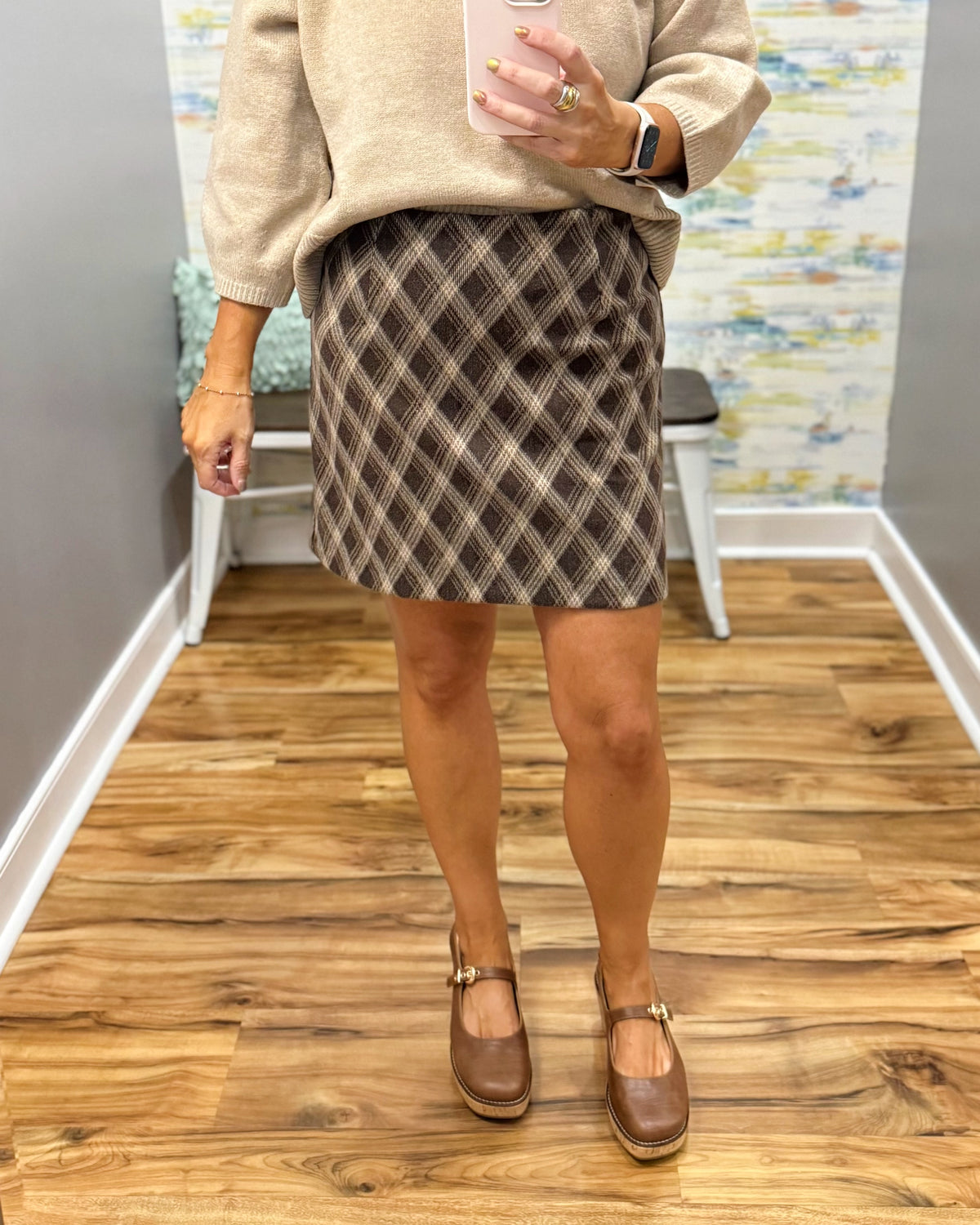 A person wearing a brown and beige argyle skirt with a beige sweater and Corkys Pity Party shoes in Cognac shoes, standing in a home interior with wood floors and a bench in the background.