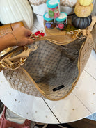 A woven beige shoulder bag with gold-tone hardware and a patterned interior lining, held open by a hand over a white wooden table.