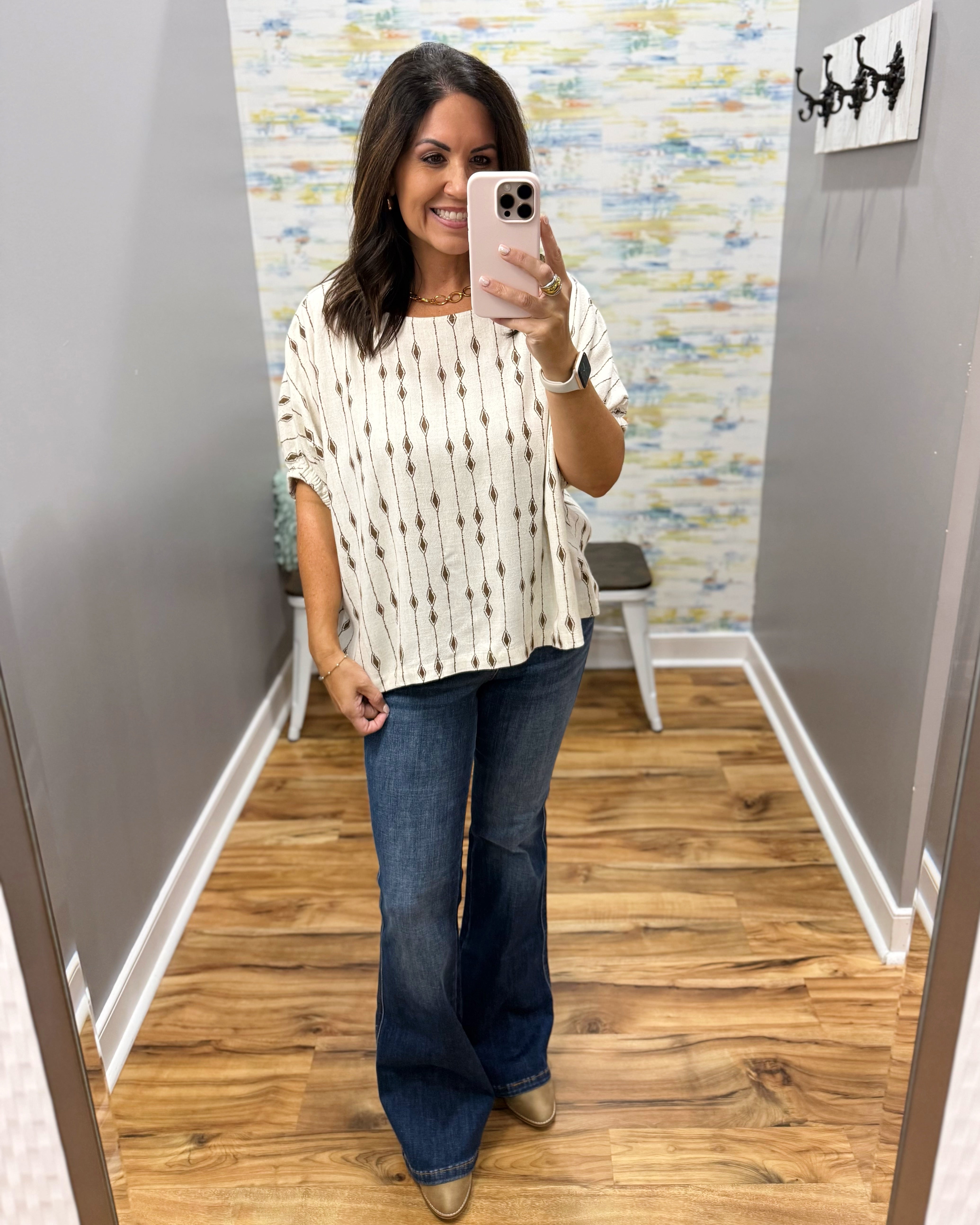 A woman takes a mirror selfie in a fitting room wearing a cream short-sleeve top with a brown ikat pattern, paired with dark blue flare jeans and tan boots.