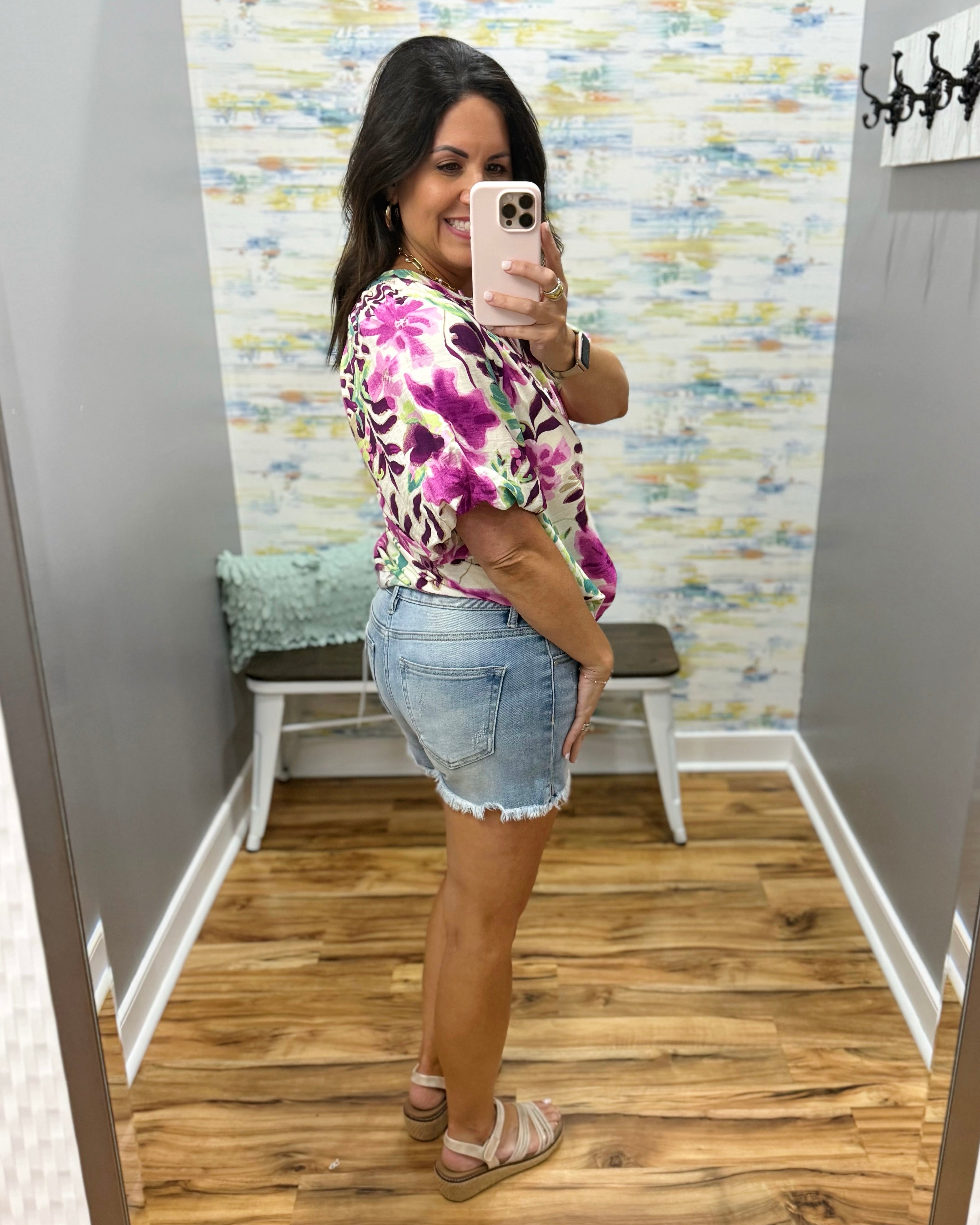 Woman taking a mirror selfie wearing a floral top and denim shorts in a room with patterned wallpaper.