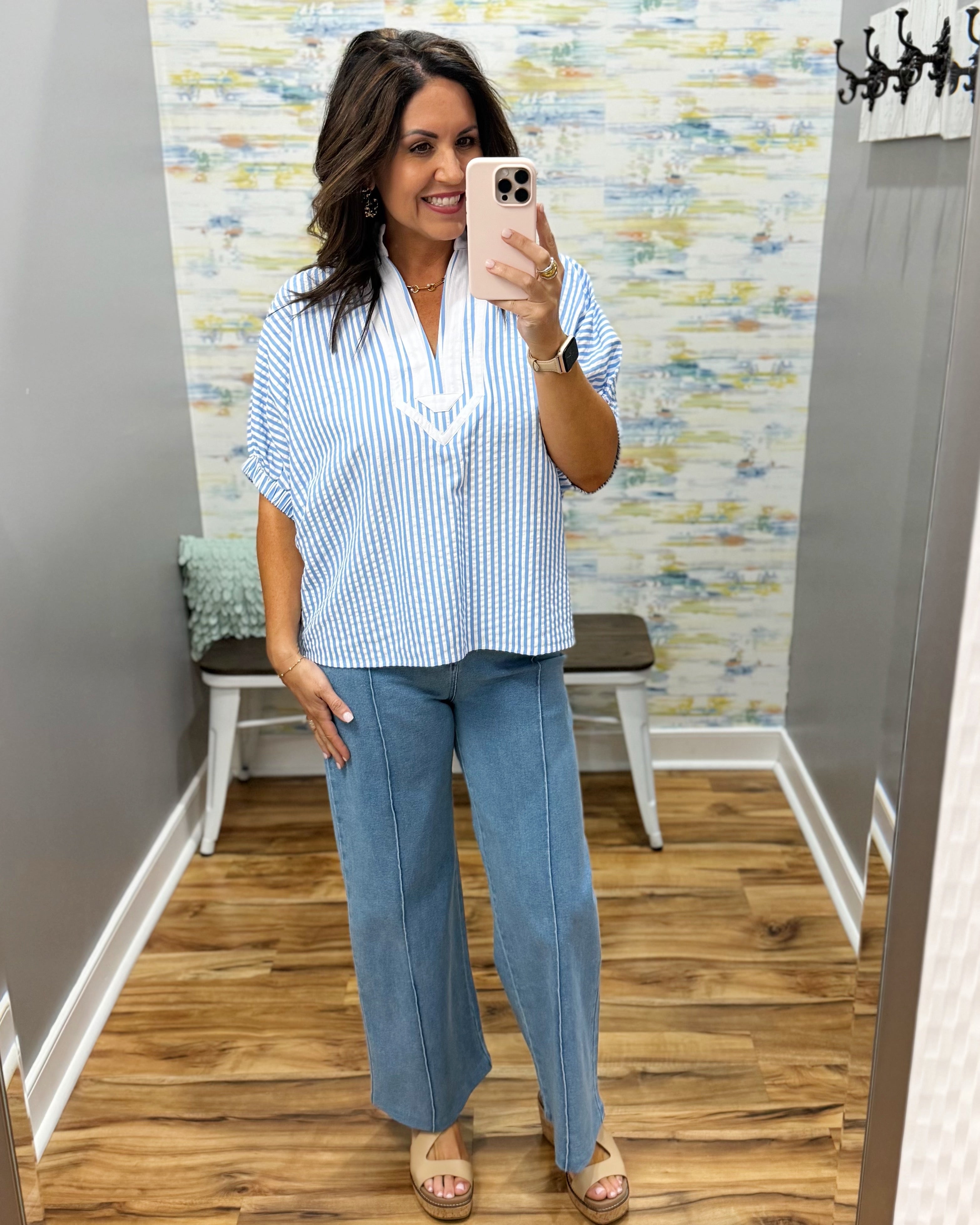 Woman taking a mirror selfie wearing a striped shirt and blue pants in a room with patterned wallpaper.