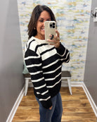 A woman takes a mirror selfie wearing a black and white horizontally striped knit sweater, blue jeans, and a light-colored phone case in a fitting room with wood floors and a patterned wall.