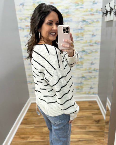 Woman taking a mirror selfie wearing a white sweater with black stripes and blue jeans.