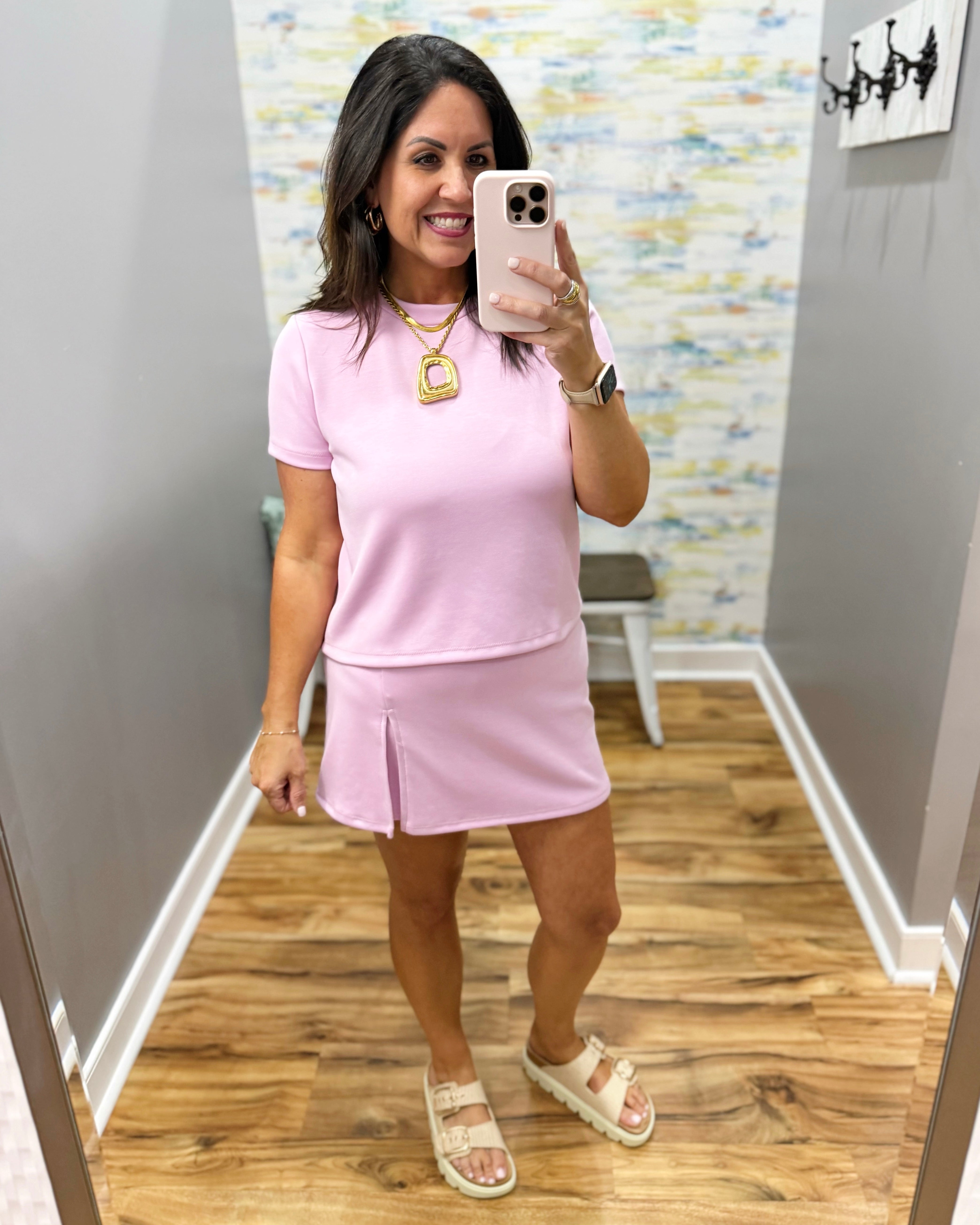 Woman taking a mirror selfie wearing a pink outfit in a room with a patterned wall.