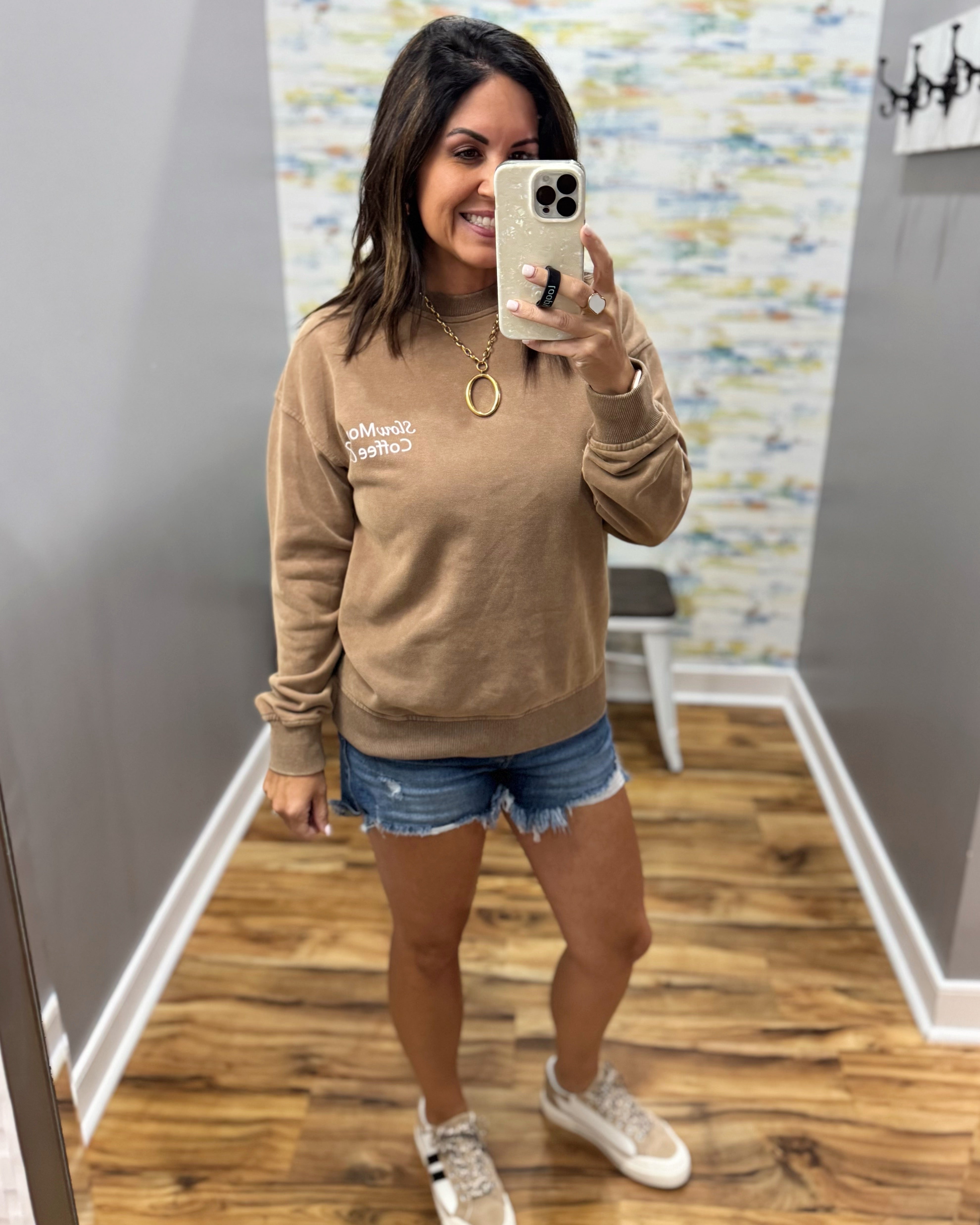 A woman takes a mirror selfie wearing a tan crewneck sweatshirt with white text on the left chest, paired with denim shorts and white sneakers in a room with wood floors and gray walls.