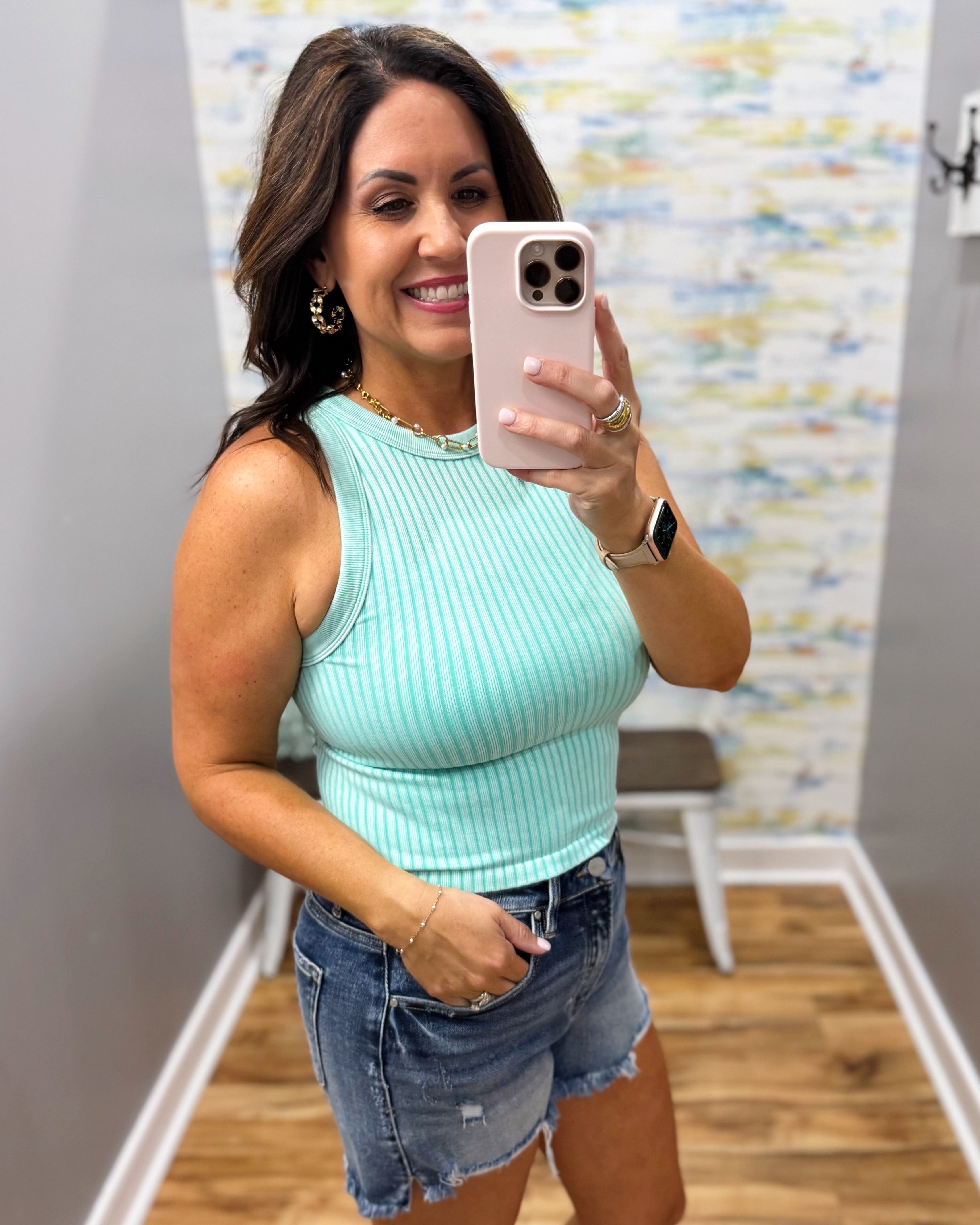 Woman taking a mirror selfie wearing a light blue ribbed tank top and denim shorts.