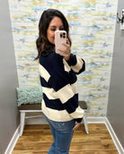 A woman takes a mirror selfie wearing a navy blue and cream striped sweater, blue jeans, and a light pink phone. The background shows a gray wall, a decorative bench with a blue-green pillow, and a colorful abstract wallpaper.