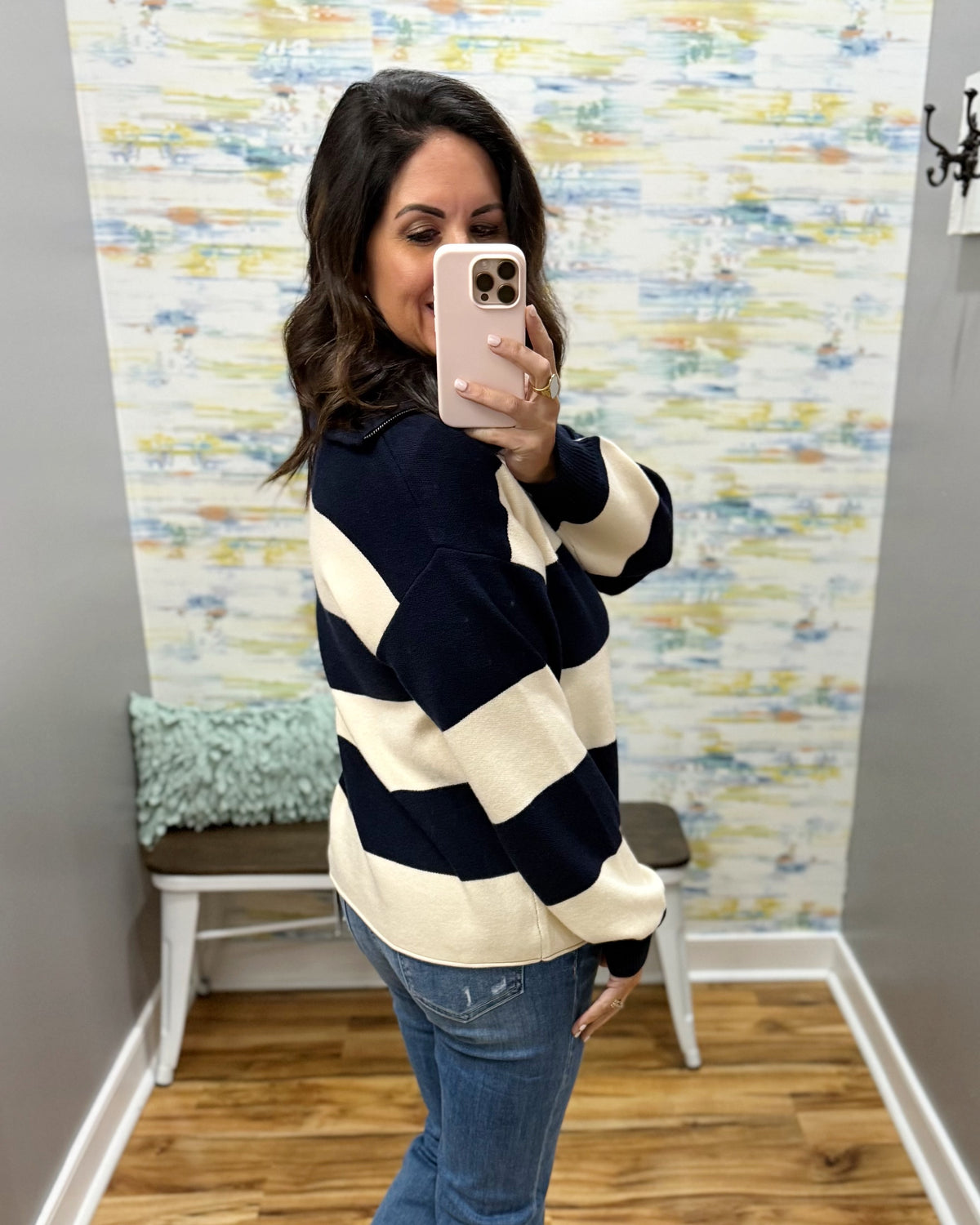 A woman takes a mirror selfie wearing a navy blue and cream striped sweater, blue jeans, and a light pink phone. The background shows a gray wall, a decorative bench with a blue-green pillow, and a colorful abstract wallpaper.