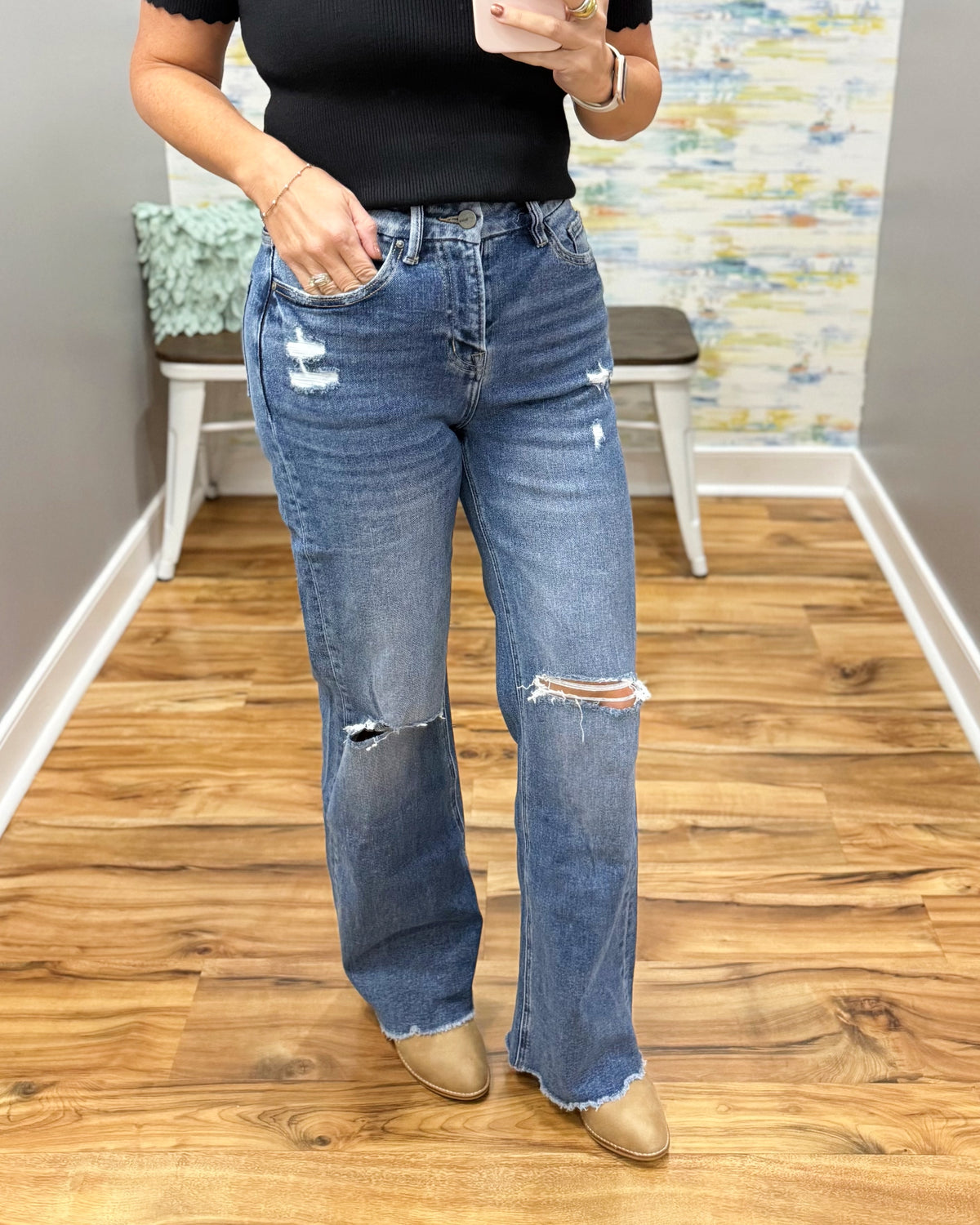 A person wearing mica wide leg distressed jeans with ripped details on the thighs and knees, paired with a black top and tan boots, standing in an indoor room with wood floors and a bench.