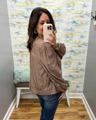 A woman takes a mirror selfie wearing a taupe brown satin bomber jacket and blue jeans in a room with patterned wallpaper and a wooden floor.
