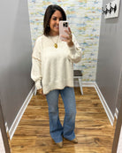A woman takes a mirror selfie in a fitting room wearing a cream-colored, oversized long-sleeve knit sweater, blue flared jeans, and beige pointed-toe flats.