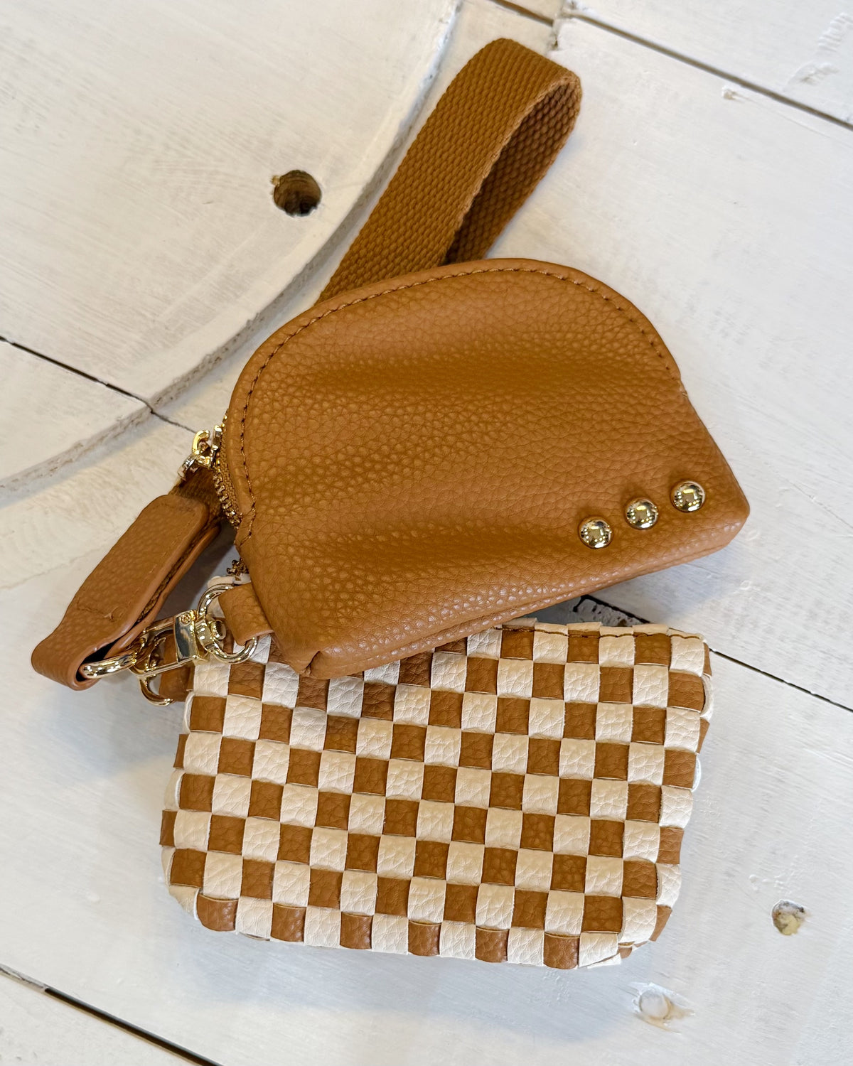 Two small leather wristlet or coin purse-style accessories on a white painted wood surface. The top piece is smooth tan leather with a wrist strap and three gold-tone rivet details.