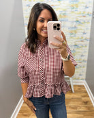 A woman taking a mirror selfie wearing a red and white gingham button-down shirt with puff sleeves and a ruffled hem, paired with blue jeans.