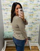 A woman takes a mirror selfie in a fitting room wearing a brown and beige horizontally striped long-sleeve top and blue jeans.