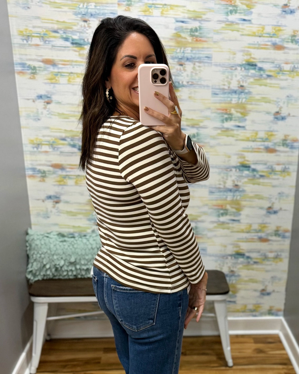 A woman takes a mirror selfie in a fitting room wearing a brown and beige horizontally striped long-sleeve top and blue jeans.