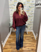 A woman takes a mirror selfie wearing a long-sleeve maroon satin blouse with puffed sleeves and blue flare jeans in a fitting room.