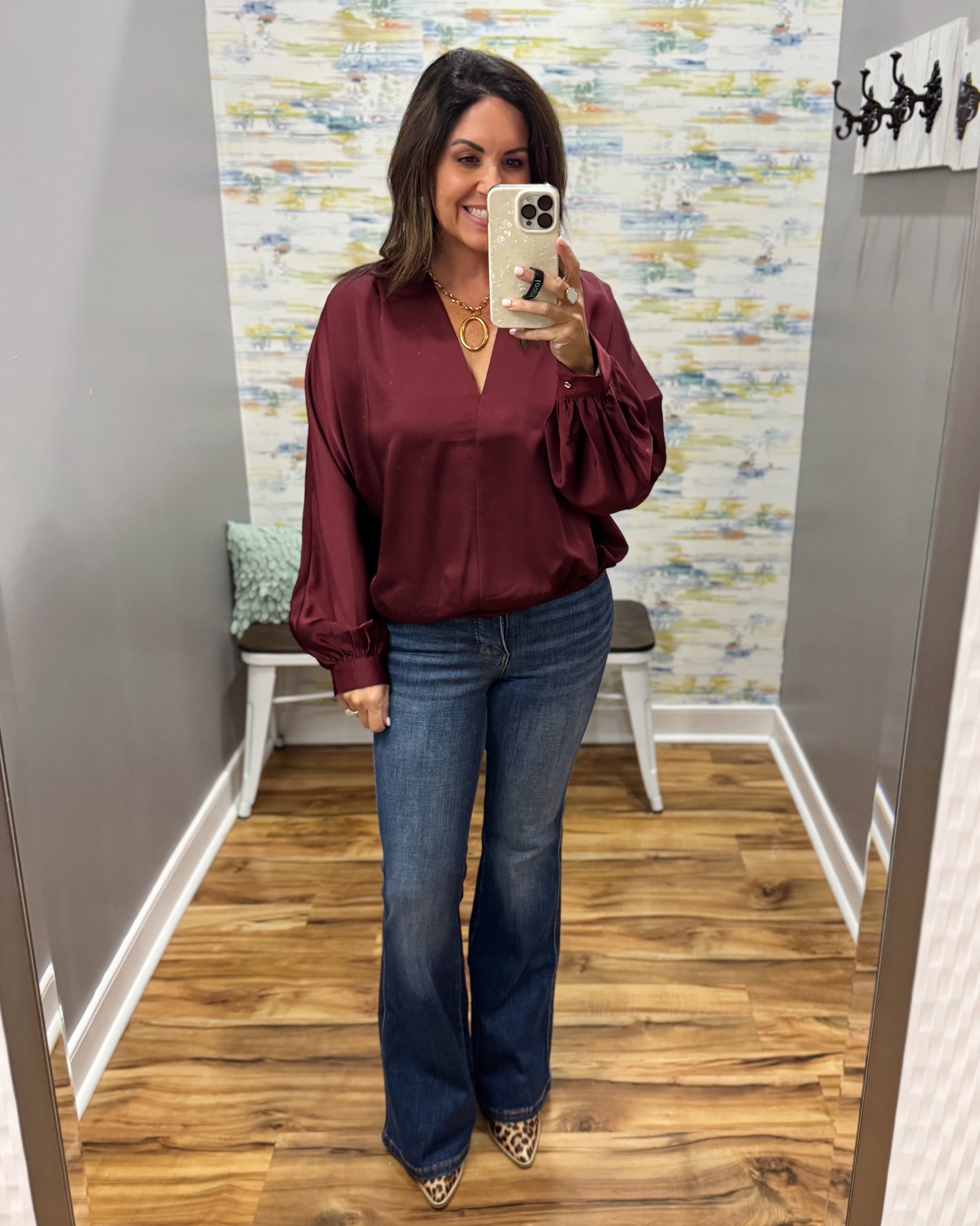 A woman takes a mirror selfie wearing a long-sleeve maroon satin blouse with puffed sleeves and blue flare jeans in a fitting room.