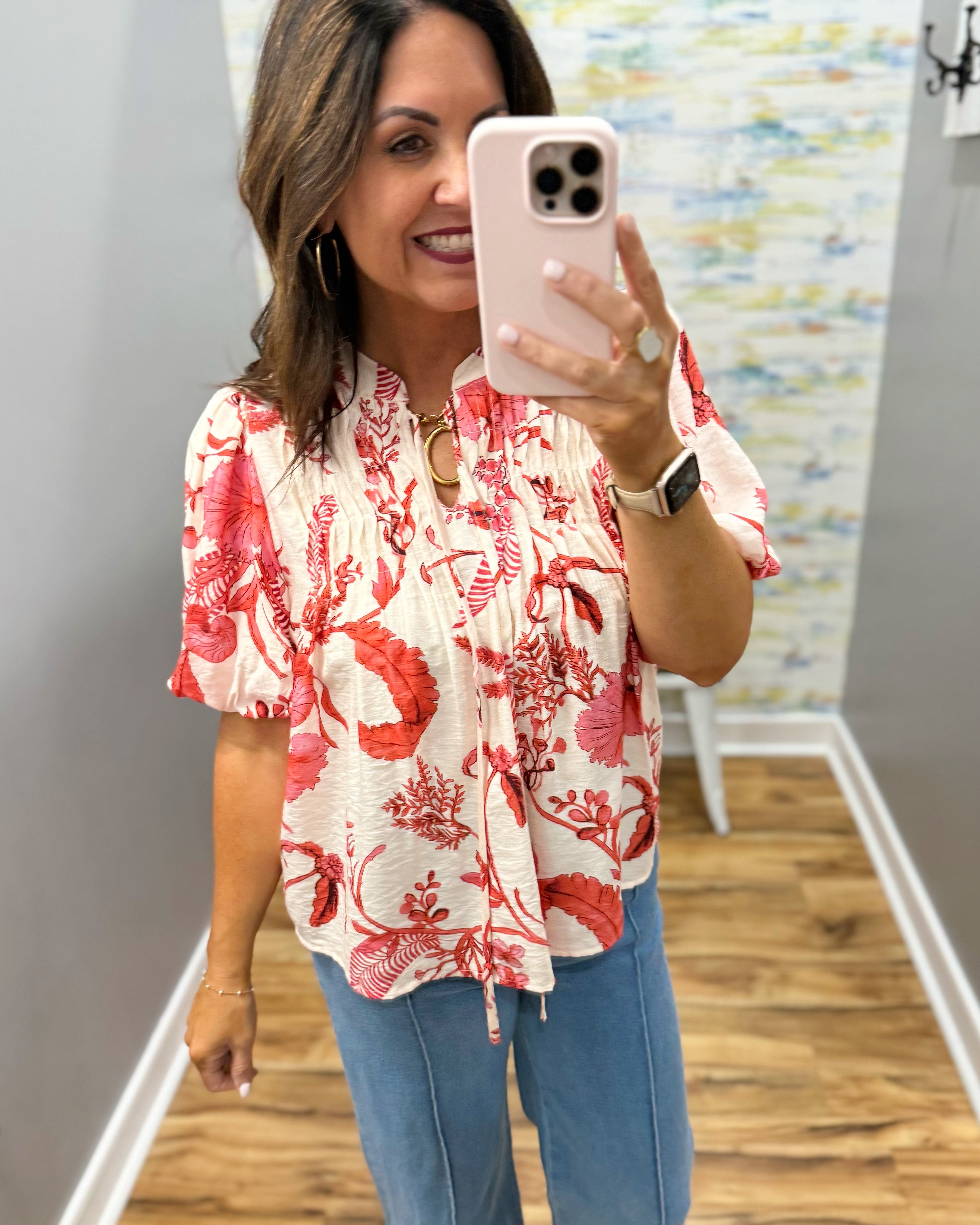 Woman taking a mirror selfie wearing a red floral blouse and blue jeans.