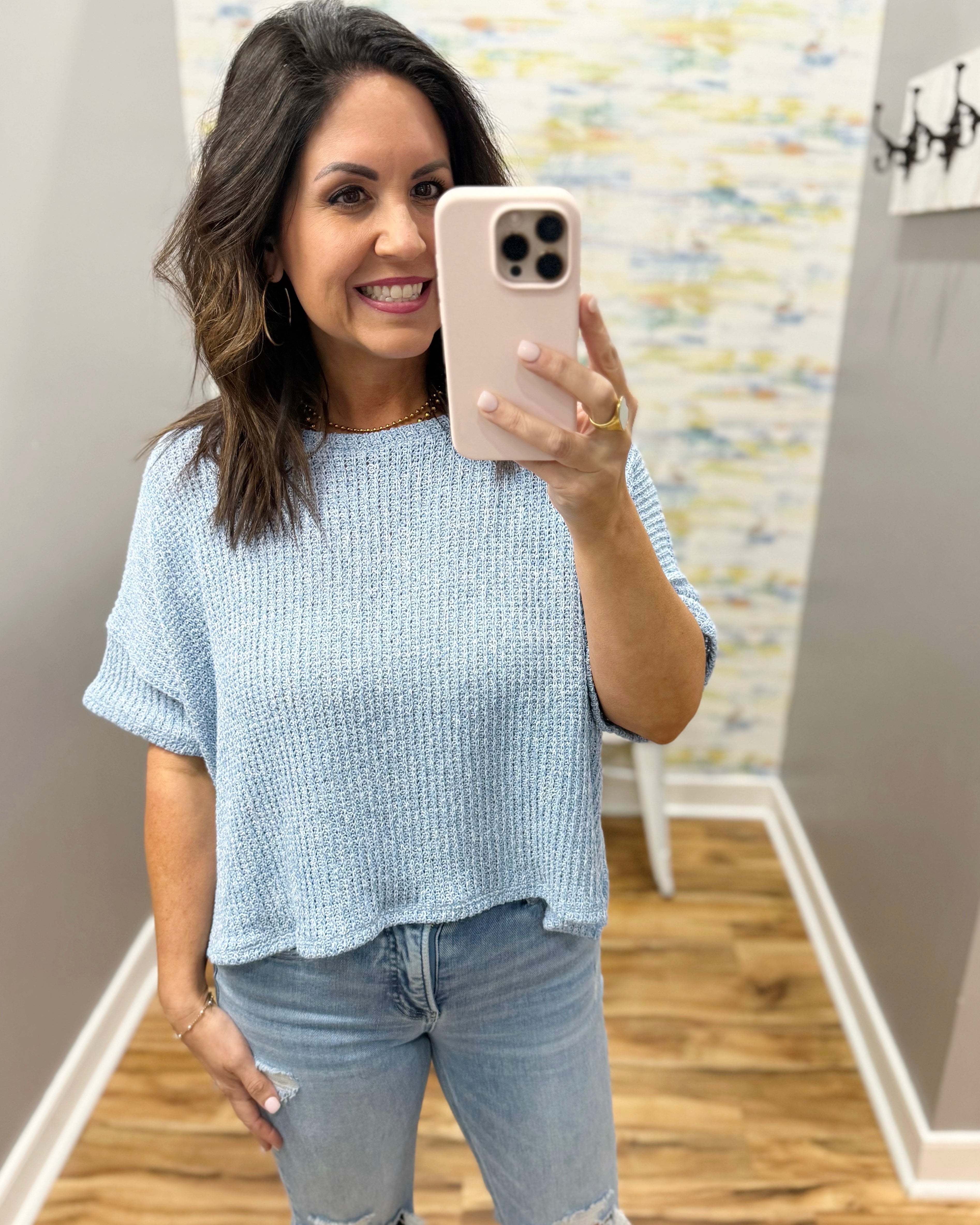 Woman taking a mirror selfie wearing a light blue top and jeans in a room with patterned wallpaper.