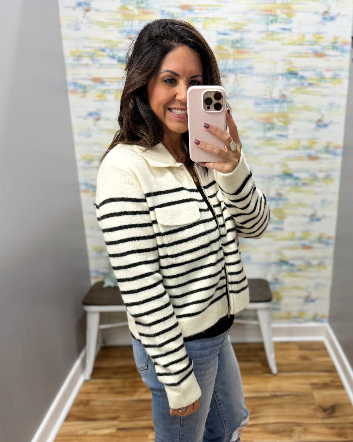 A woman taking a mirror selfie in a fitting room wearing a cream and black horizontally striped knit zip-up jacket with a collar and chest pocket, paired with blue jeans.