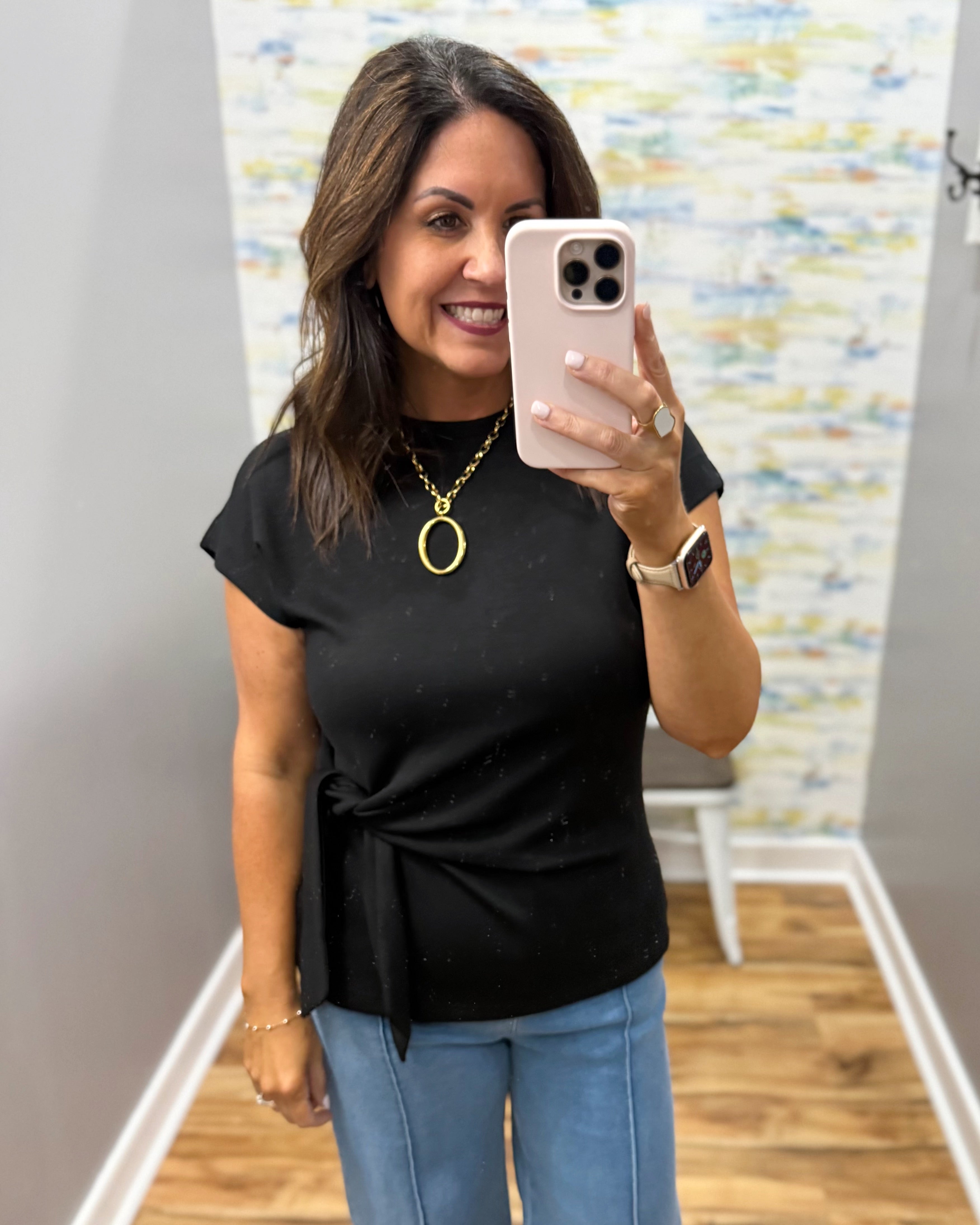 Woman taking a mirror selfie wearing a black top and blue jeans, holding a phone.