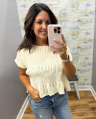 A woman taking a mirror selfie wearing a cream-colored short-sleeve smocked blouse and blue jeans in an indoor setting.