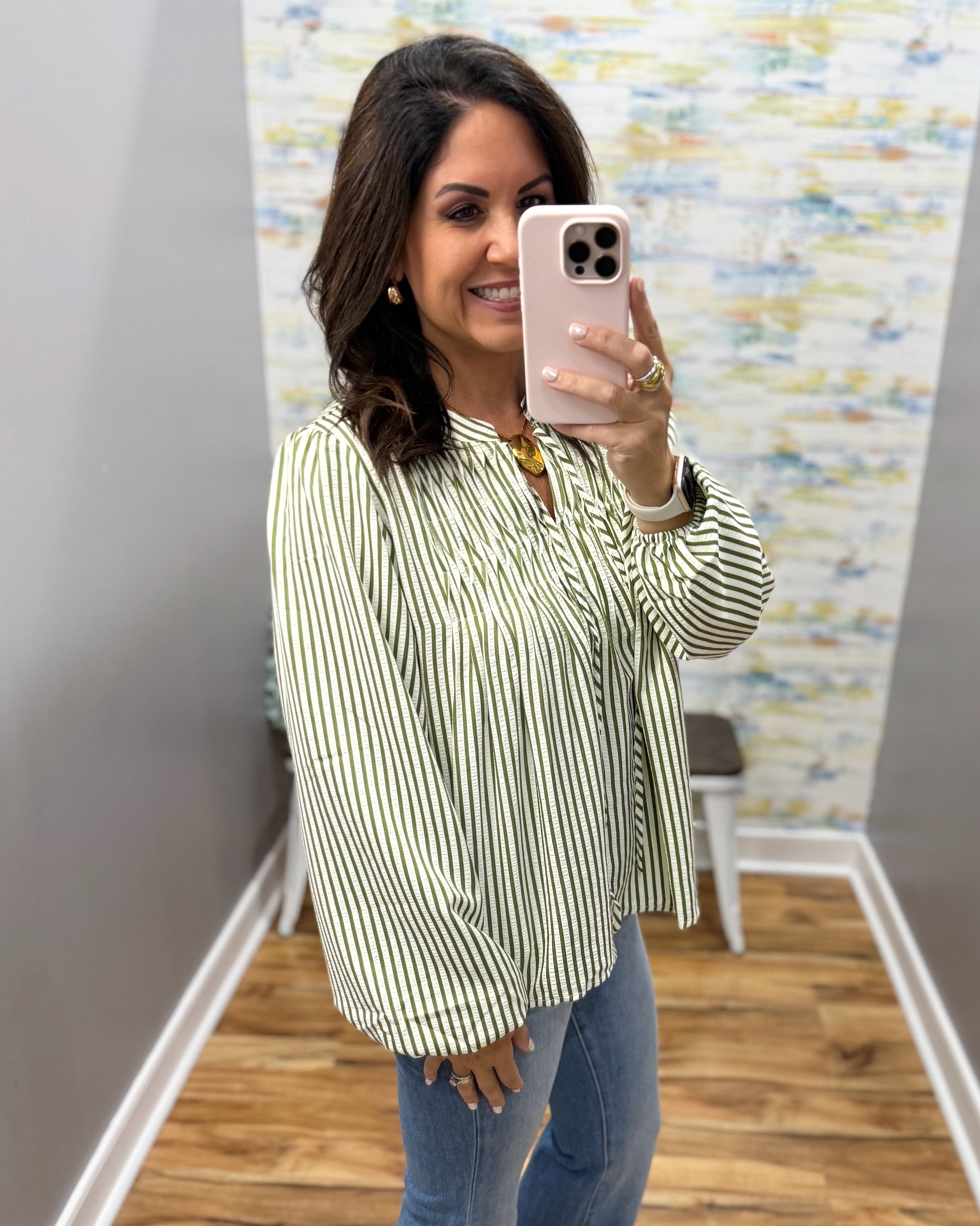 A woman takes a mirror selfie wearing a green and white vertically striped long-sleeve blouse with blue jeans, holding a light pink smartphone.