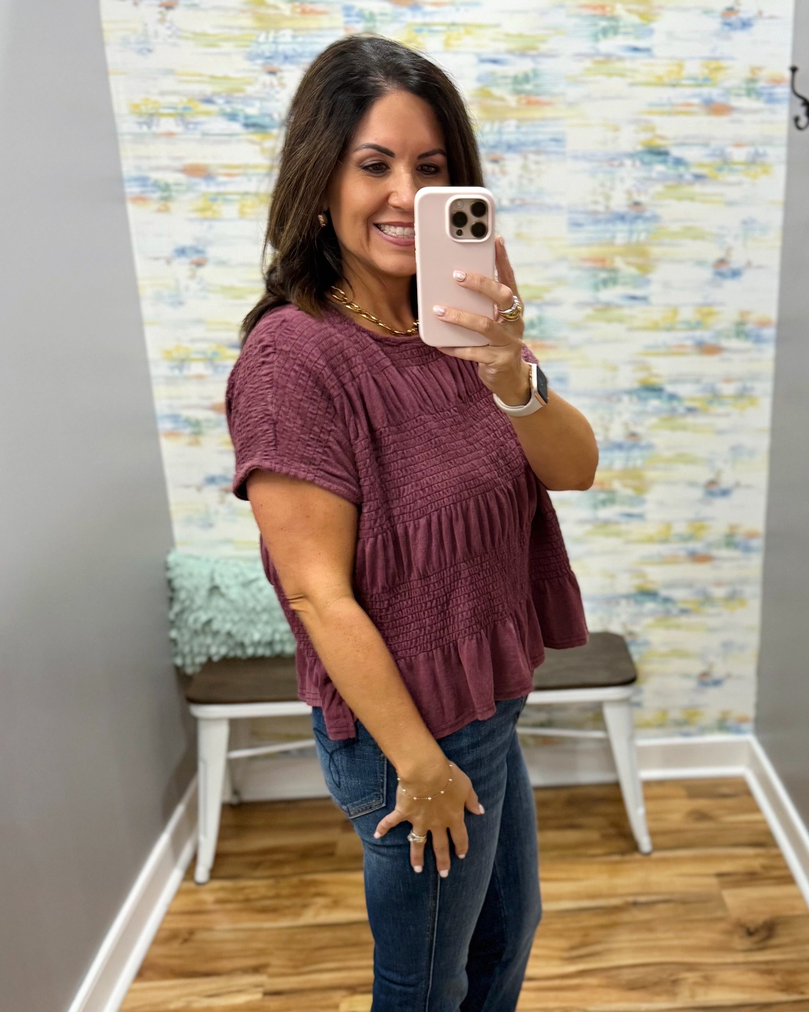 A woman takes a mirror selfie wearing a short-sleeve, smocked, tiered peplum top in a dusty mauve-purple color, paired with blue jeans. The setting is a fitting room with a patterned wallpaper background and a wooden floor.