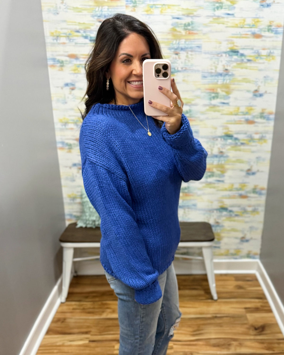 A woman takes a mirror selfie wearing a vibrant royal blue textured knit sweater and light-wash jeans in a home setting with a patterned wallpaper background.