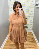 A woman takes a mirror selfie wearing a short-sleeve button-up shirt dress in a warm tan or camel color. The dress features a collar, chest pocket, and tiered ruffle hem.