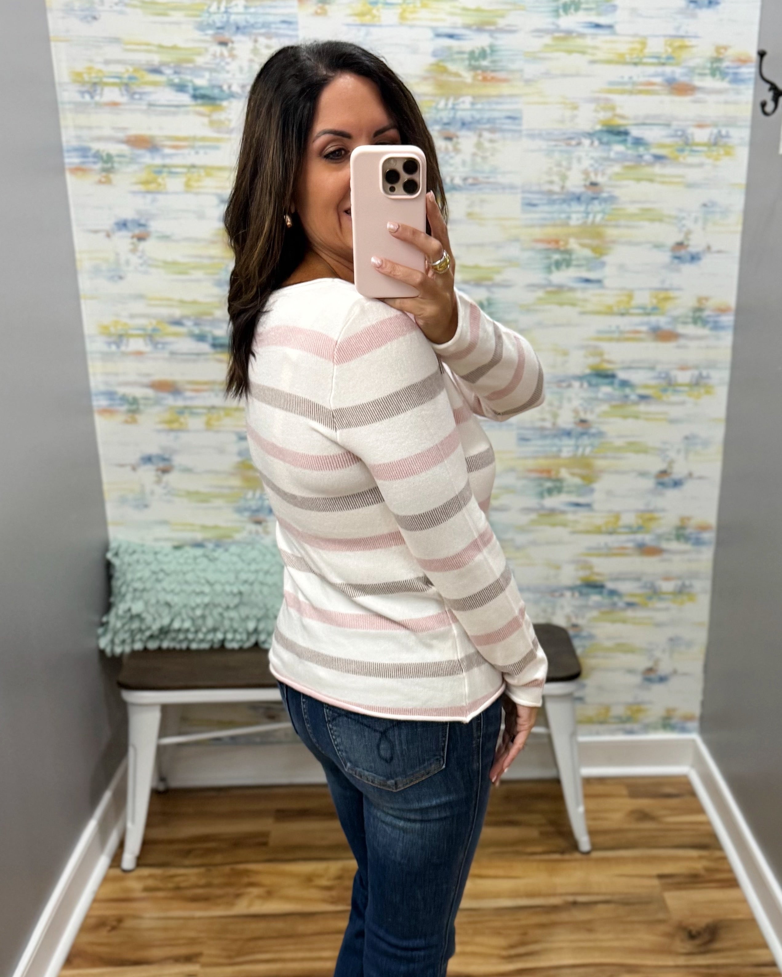 A woman takes a mirror selfie wearing a long-sleeve knit top with horizontal stripes in white, light pink, and taupe, paired with dark blue jeans.